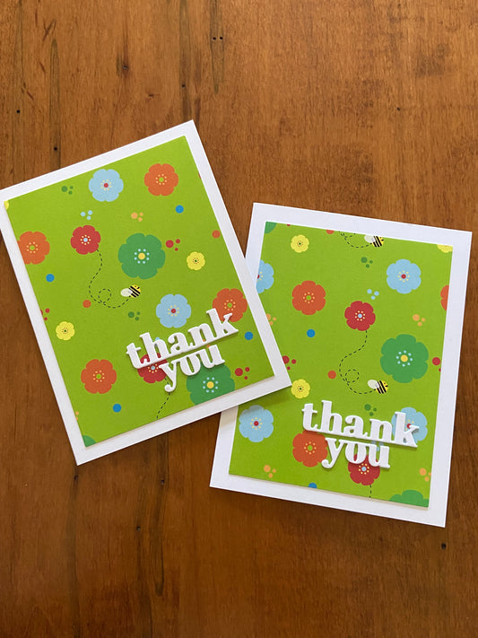 2 thank you cards. A lime green background with yellow, green, blue and orange flowers scattered through out. A few bumble bees are flying amongst the blooms. A bright with thanks you die cut sentiment is placed on the lower right corner of the card.