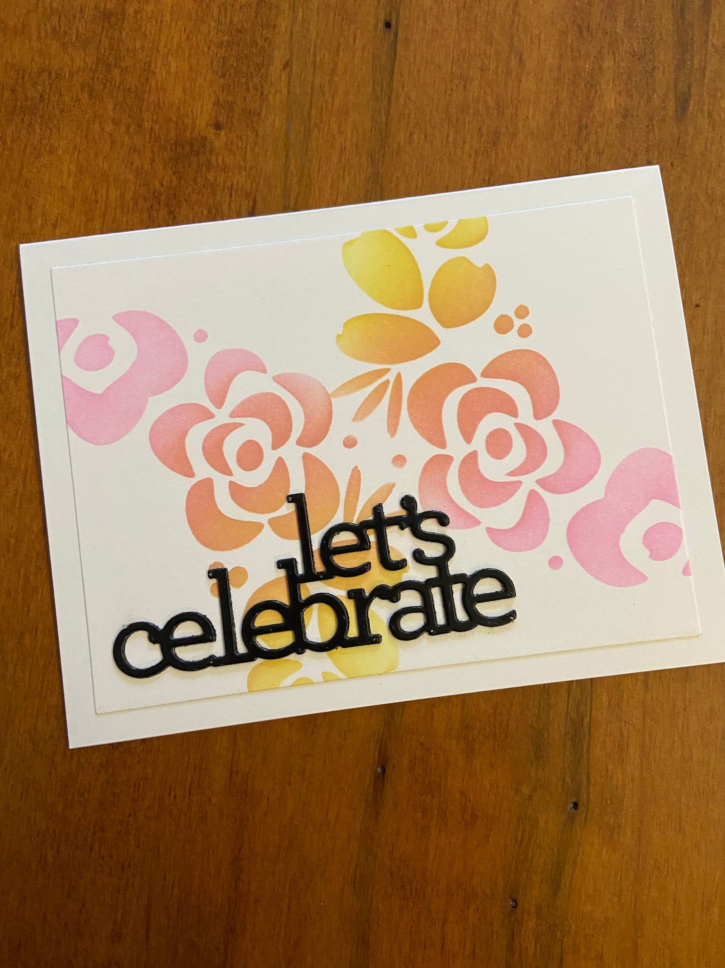 Modern Flowers Let’s Celebrate handmade card. Ink blended modern flowers stencil in shades of pink, orange and yellow. A lets celebrate die cut sentiment in black to finish in bottom left corner.