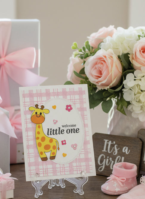 Baby shower card with a giraffe illustration and 'welcome little one' text, placed on a stand with flowers and baby shoes in the background.
