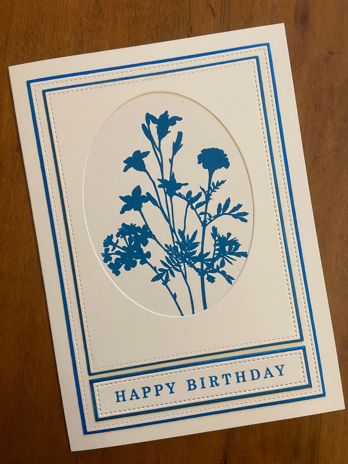 Silhouette wild flowers are stamped in a metallic marine blue ink on ivory cardstock. The 5x7 card has 2 layers of blue matted die cut panels and a happy birthday sentiment is stamped in matching blue.