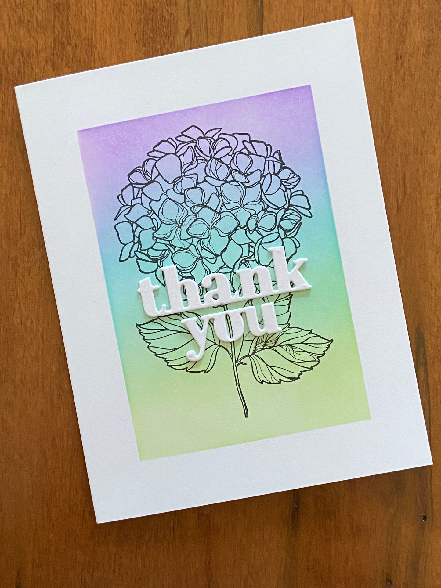 Thank you handmade card. Ink blended background in shades of green, turquoise and purple. A hydrangea cluster black outline stamp is the focal image with bold white thank you die cut sentiment on top.