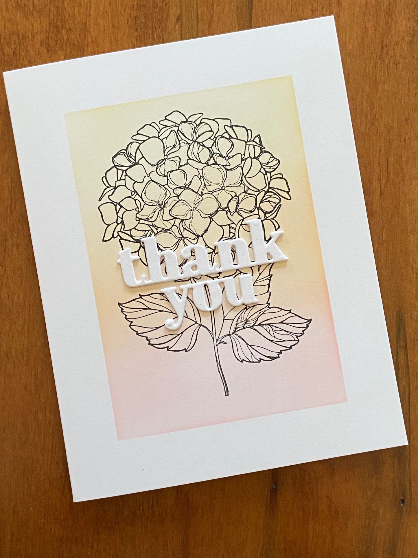 Thank you handmade card. Ink blended background in shades of pink, orange and yellow. A hydrangea cluster black outline stamp is the focal image with bold white thank you die cut sentiment on top.