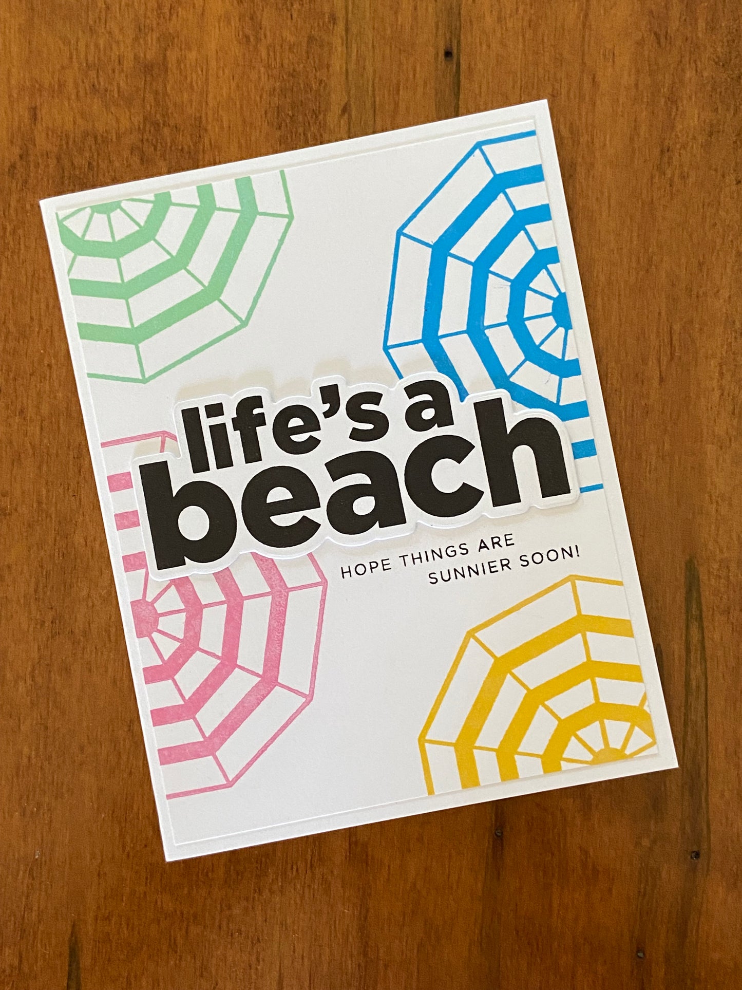 Life’s A Beach Encouragement handmade card. A white background has some top-down view green , pink, green, and yellow umbrellas and a bold black Life’s a beach sentiment centred. Hope things are sunnier soon is the sun