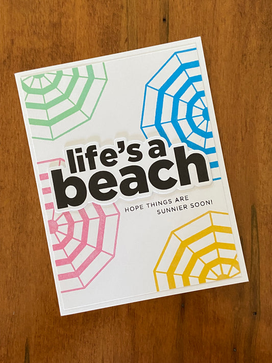 Life’s A Beach Encouragement handmade card.  A white background has some top-down view green , pink, green, and yellow umbrellas and a bold black Life’s a beach sentiment centred. Hope things are sunnier soon is the sun