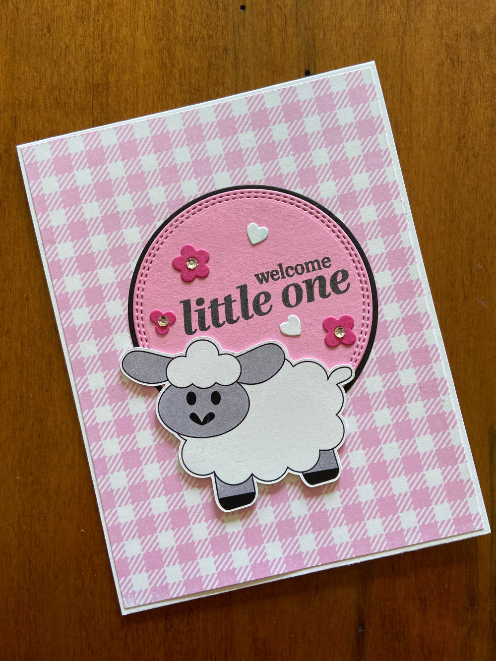 A pink and white gingham greeting card with a hand stamped sentiment 'Welcome Little One' and an illustration of a sheep, along with some pink flowers and silver sequins.