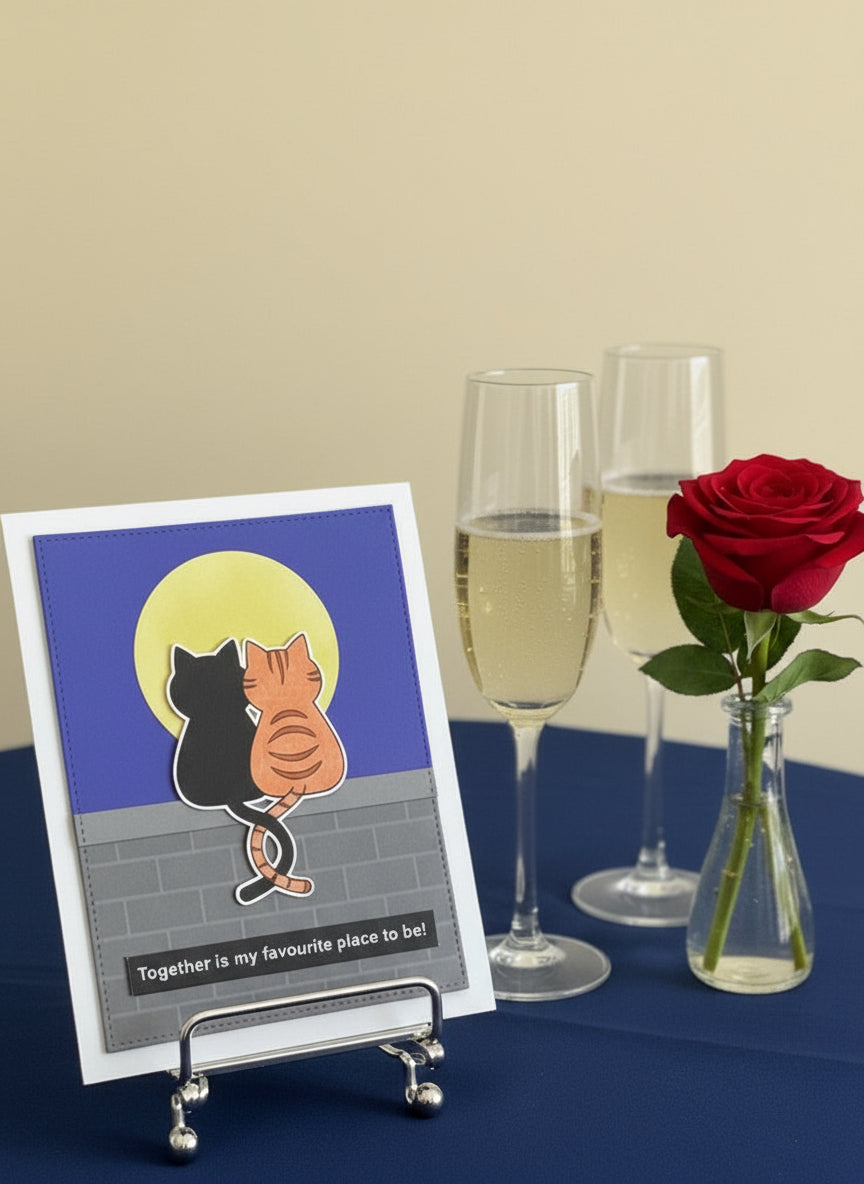Anniversary or love card with 2 cats in the moonlight on a stand next to two glasses of champagne and a rose.
