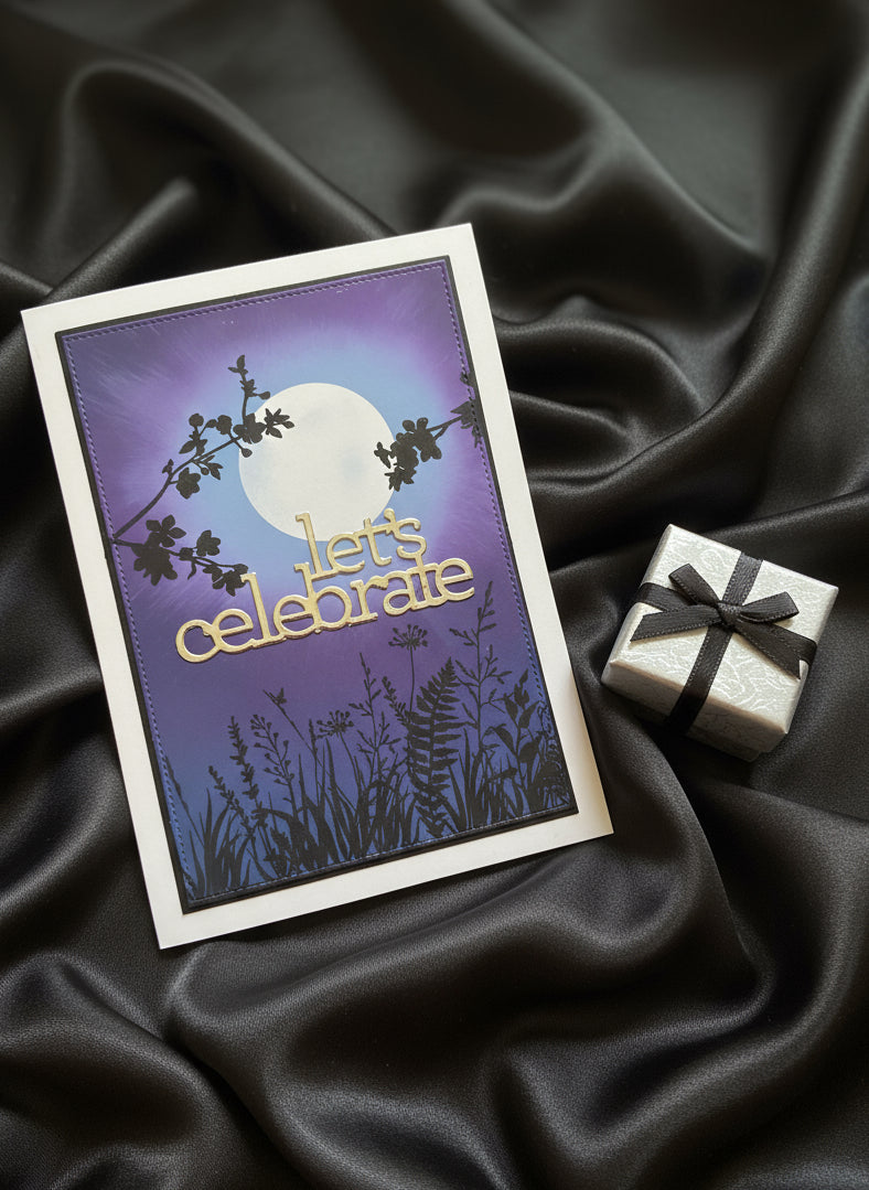 Celebration card with moonscape background and 'Let's Celebrate' sentiment and a tiny silver gift laid on black shiny fabric
