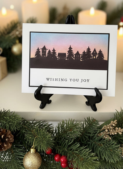 Card with silhouette of trees and villiage, 'Wishing You Joy' text, on a mantle with Christmas decorations.