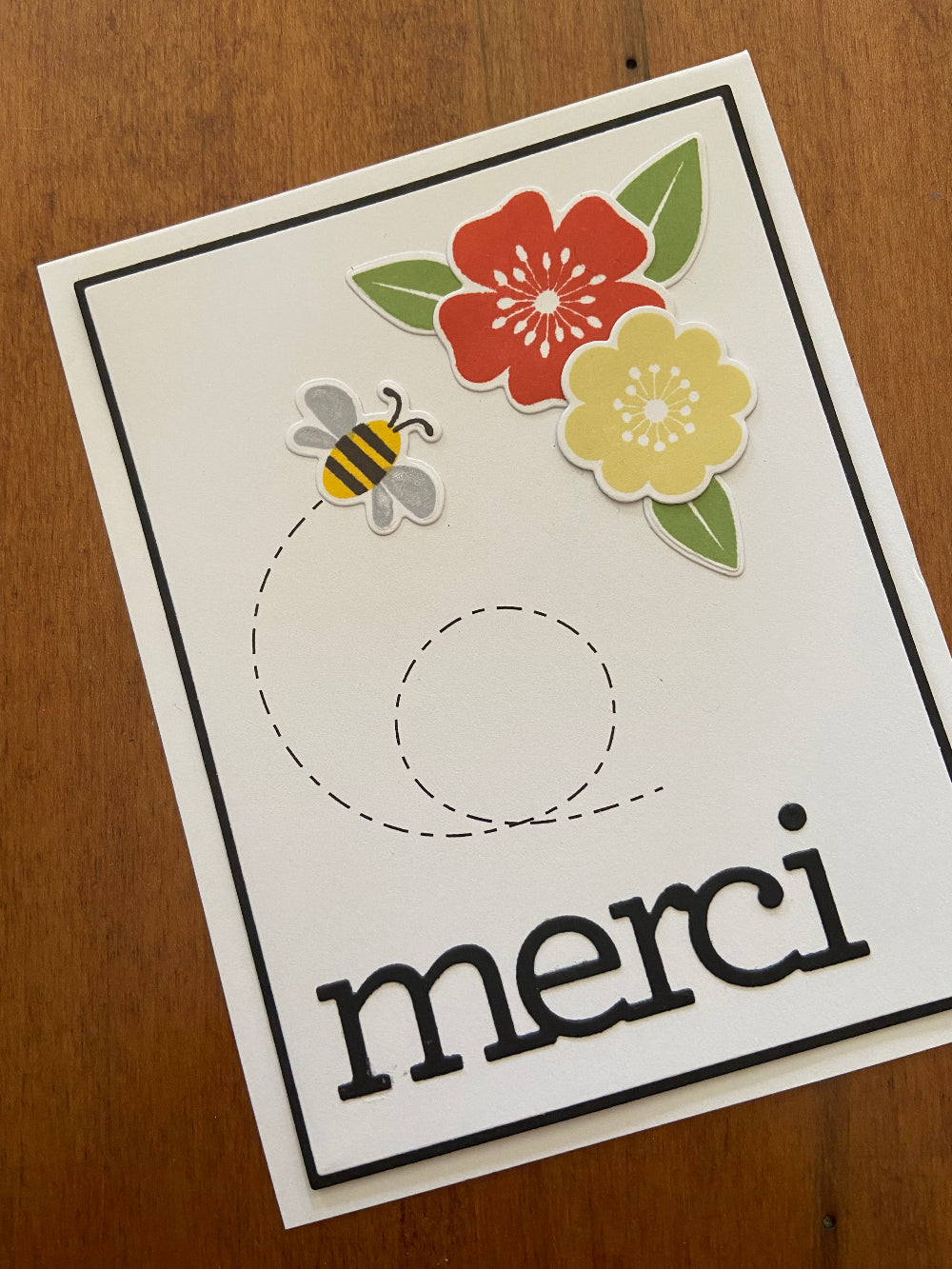 A white thank you card with 'Merci' written in black, featuring die cut floral and bee designs on the front.