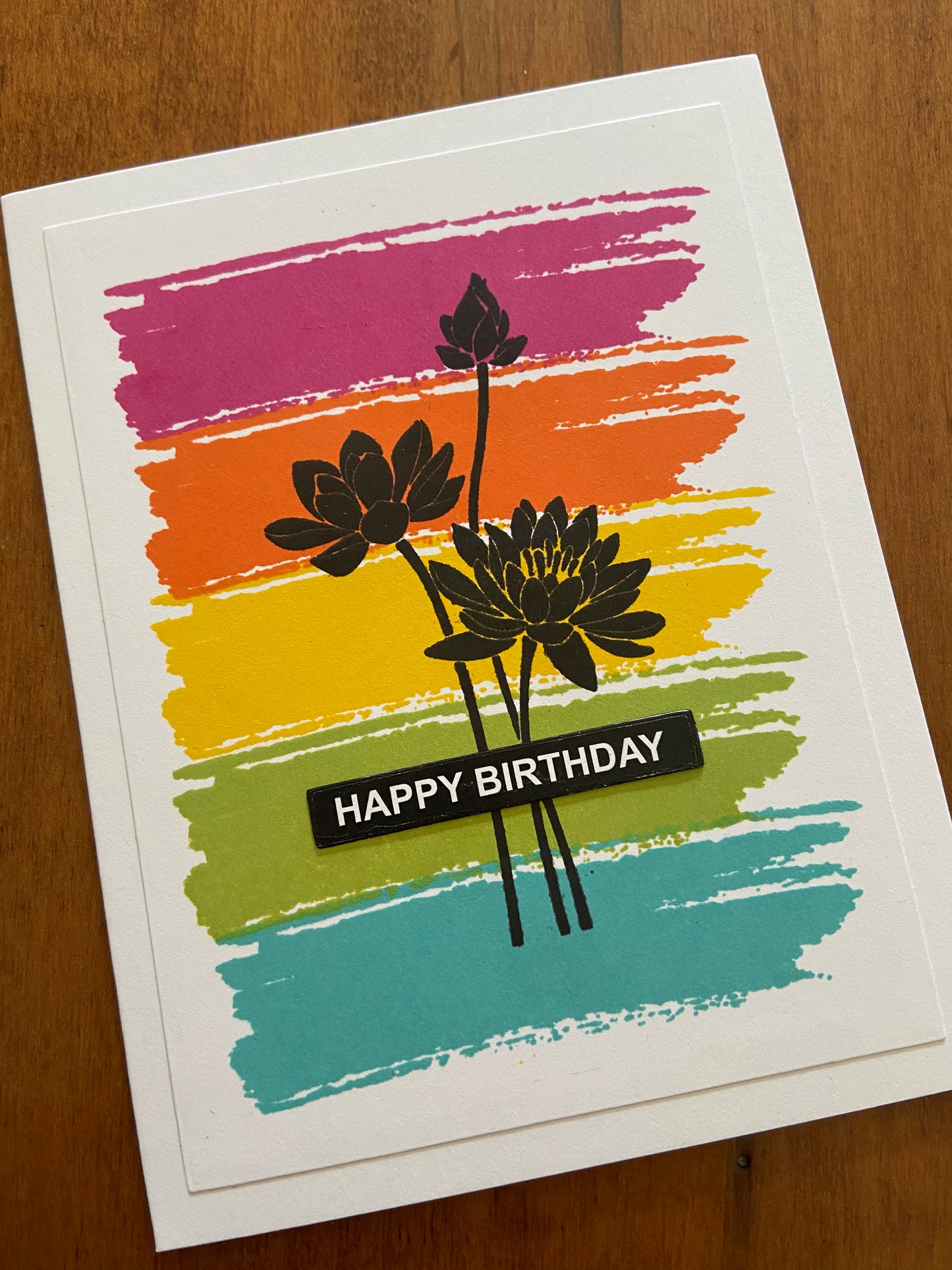 Dark pink, orange, yellow, lime green and turquoise paint stroke stamps background. A lotus silhouette stamp over top and a happy birthday sentiment over top.