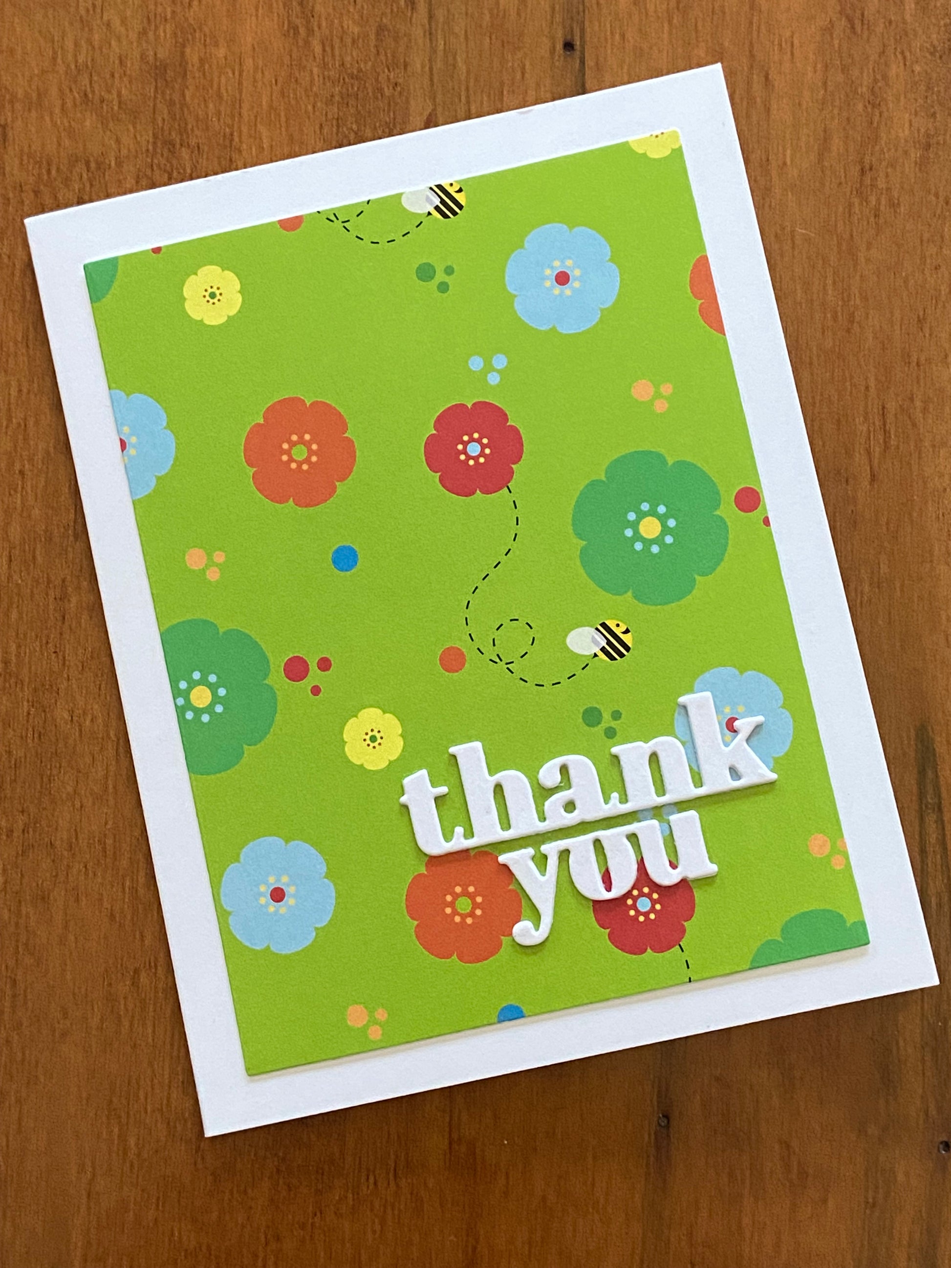 A lime green background with yellow, green, blue and orange flowers scattered through out. A few bumble bees are flying amongst the blooms. A bright with thanks you die cut sentiment is placed on the lower right corner of the card. 