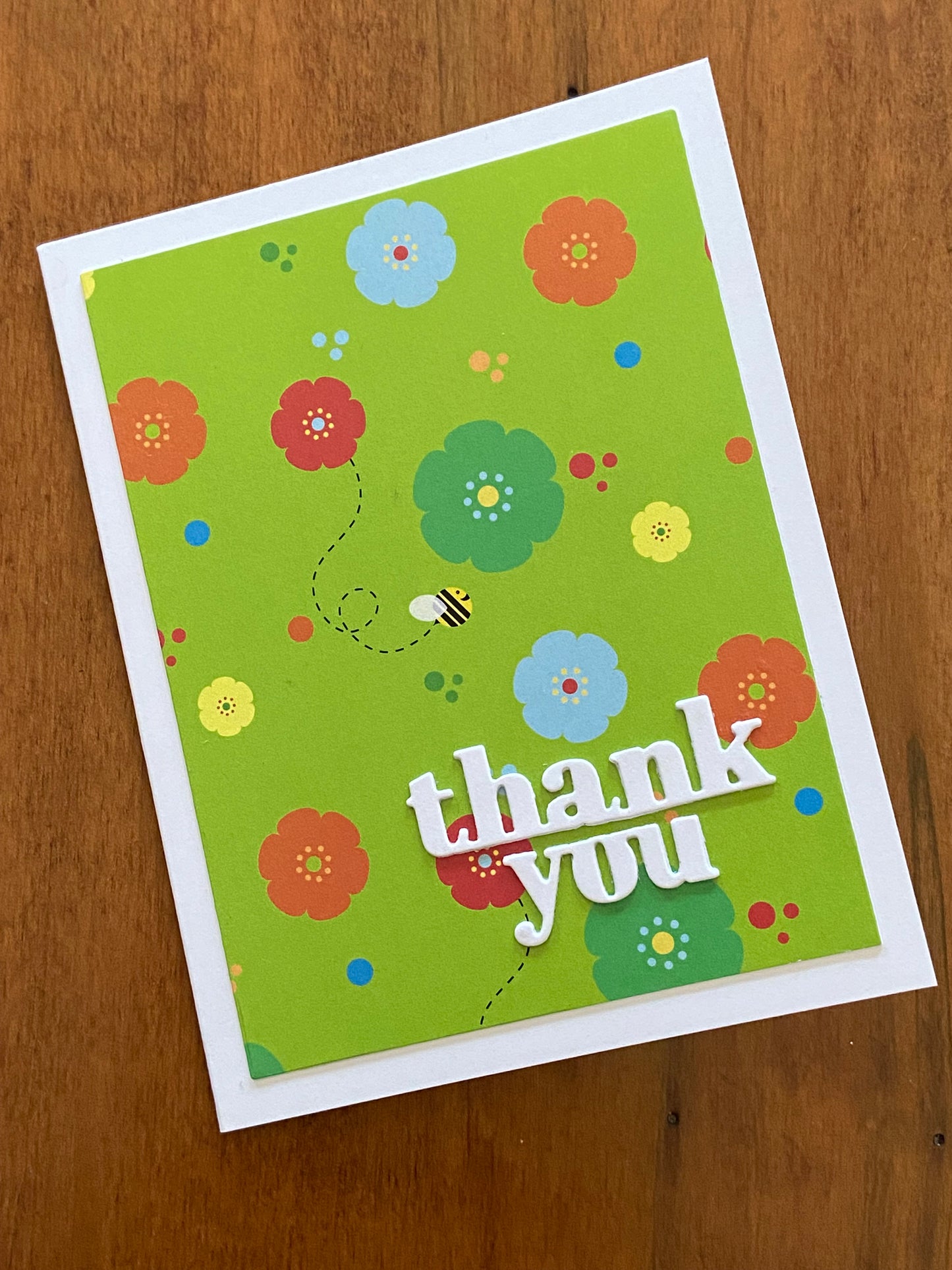 A lime green background with yellow, green, blue and orange flowers scattered through out. A few bumble bees are flying amongst the blooms. A bright with thanks you die cut sentiment is placed on the lower right corner of the card. 