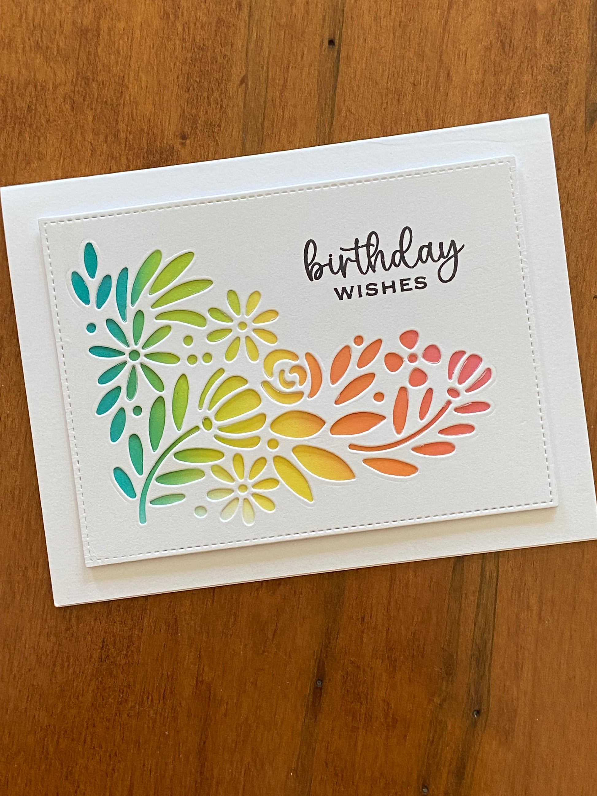 Birthday Wishes handmade card. A corner floral die cut panel is placed over another rainbow coloured ink blended panel so the the rainbow colours peek through. An elegant “Birthday Wishes” sentiment is stamped in black.