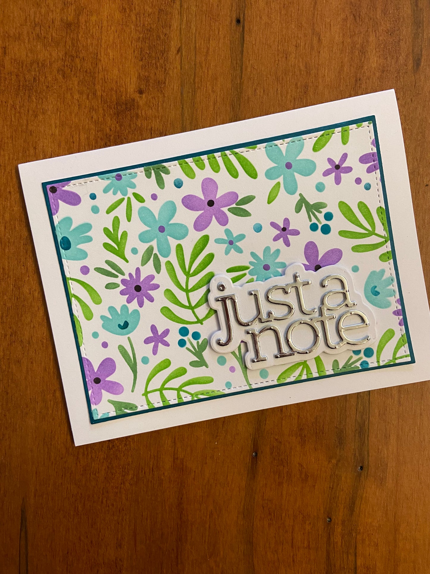 Dainty flowers and foliage just a note hand made card. Featured colours of purple, turquoise, and green. A silver metallic just a note die cut in bottom right corner finishes the card. 