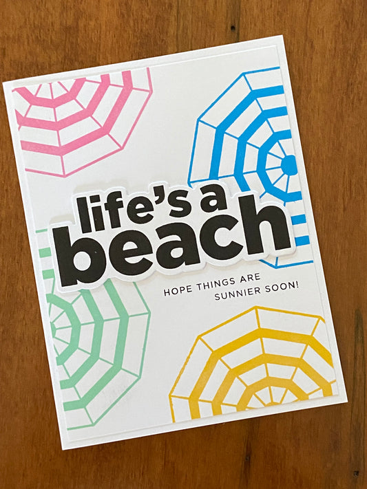 Life’s A Beach Encouragement handmade card.  A white background has some top-down view pink, blue, green, and yellow umbrellas and a bold black Life’s a beach sentiment centred. Hope things are sunnier soon is the sun sentiment. 