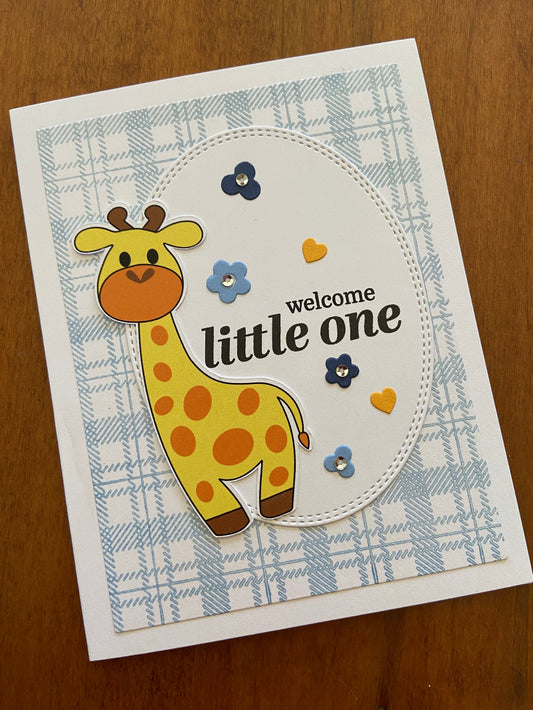 A handmade greeting card with a blue plaid background, featuring a giraffe illustration, and the text 'Welcome Little One'.