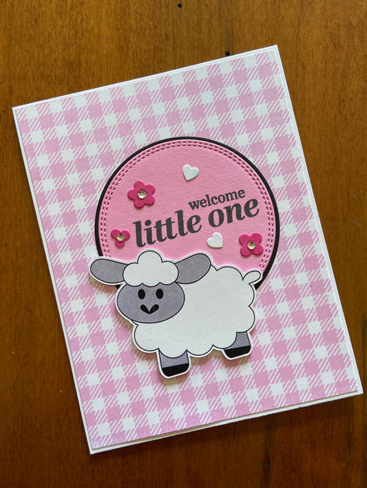 A pink and white gingham greeting card with a hand stamped sentiment 'Welcome Little One' and an illustration of a sheep, along with some pink flowers and silver sequins.