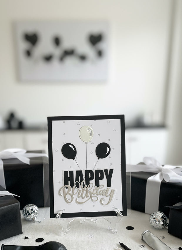Happy Birthday card with black and silver balloons on a desk with gifts and decorations.