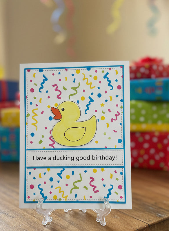 Birthday card with a rubber duck original artwork design and a confetti background; birthday gifts in the background