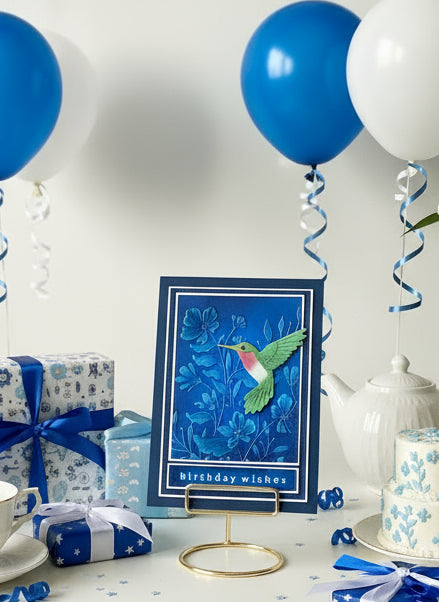 Decorative birthday card with blue floral design and green bird, surrounded by blue and white balloons and gifts.