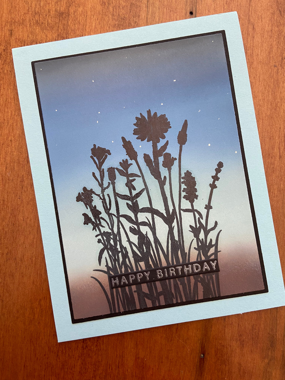 Handmade birthday card with a silhouette of wildflowers against a grey, blue, and green sky background, with 'HAPPY BIRTHDAY' text.