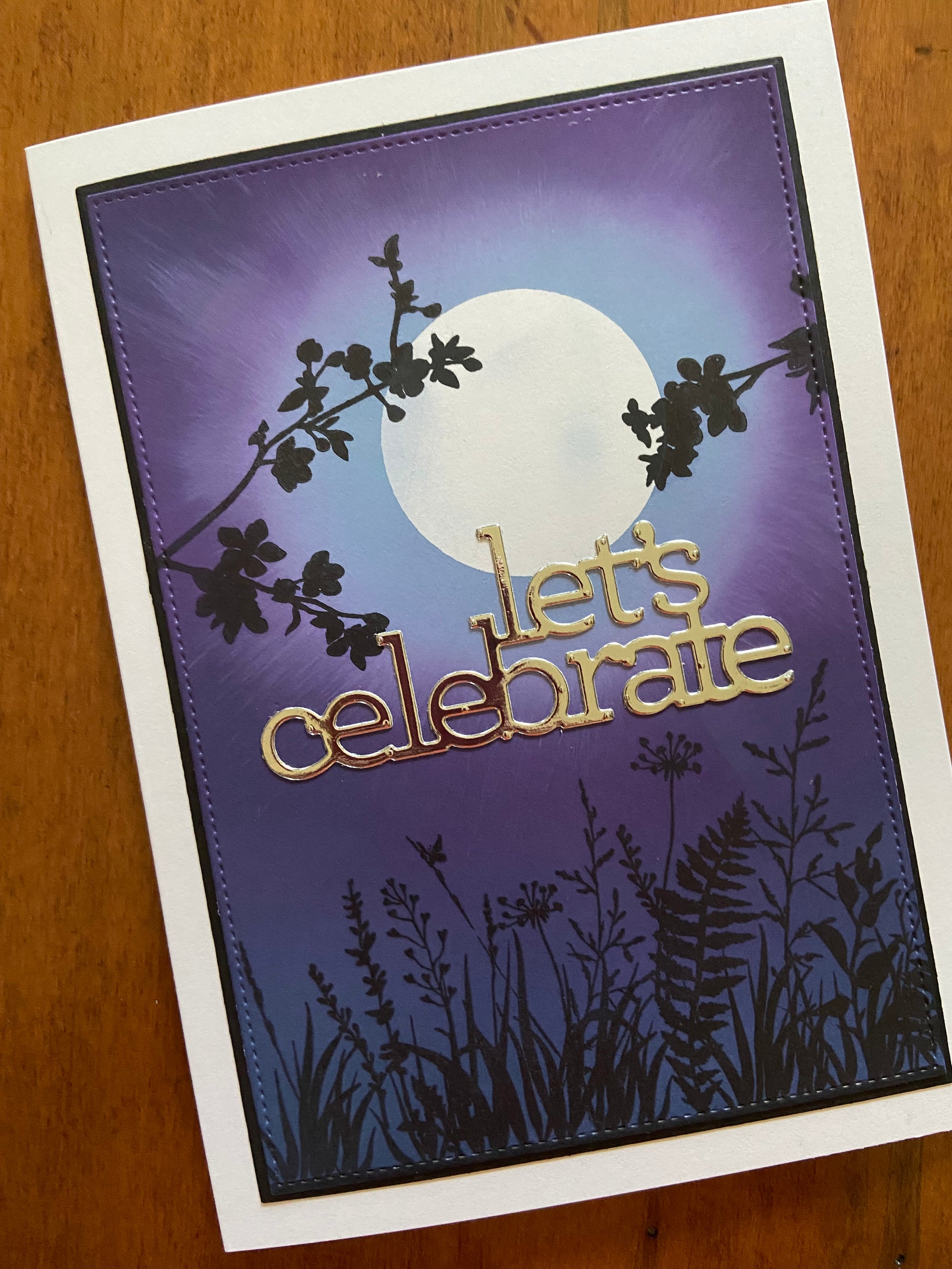 Handmade celebration card with ink-blended moonscape background, black silhouette branches and foliage, and metallic Let's Celebrate die-cut sentiment