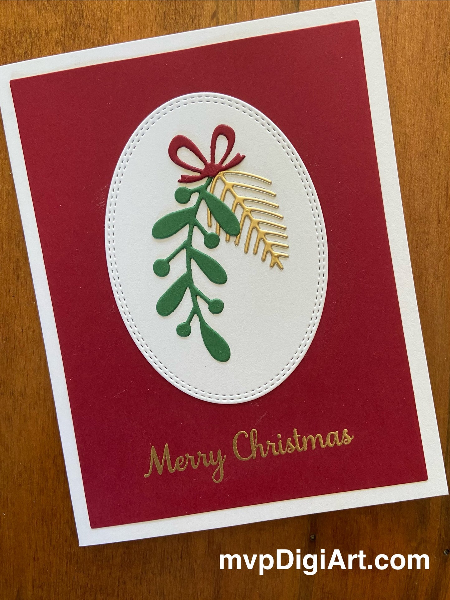 Handmade Christmas card with greenery die cuts and spruce sprigs tied with a festive bow on a stitched oval backdrop, with 'Merry Christmas' text in gold on a red background.