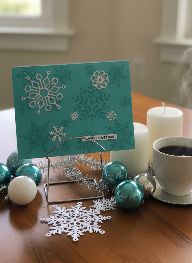 Teal Christmas card with snowflakes on a table with ornaments and a cup of coffee.