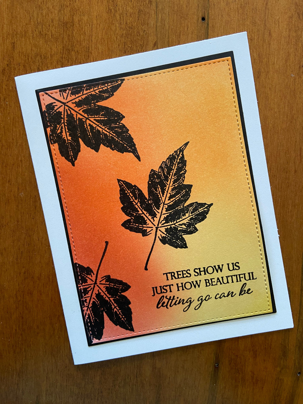 A handmade encouragement card with an autumn theme, featuring a vibrantly colored background, stamped silhouettes of maple leaves, and a message on the front. The card is placed against a wooden background.
