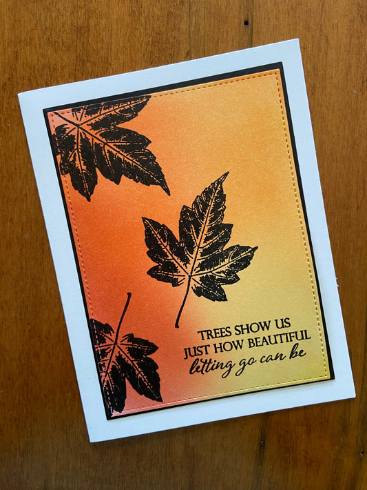 A handmade encouragement card with an autumn theme, featuring a vibrantly colored background, stamped silhouettes of maple leaves, and a message on the front. The card is placed against a wooden background.