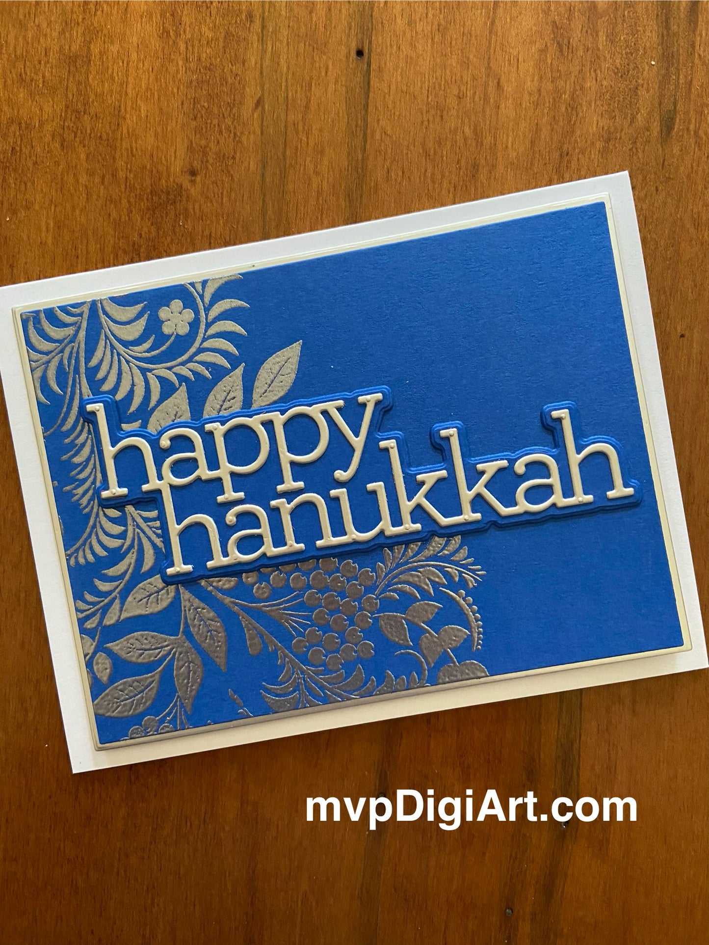 A handmade Hanukkah card with a blue background, silver vine with leaves and berrie, the text 'happy hanukkah' in silver.
