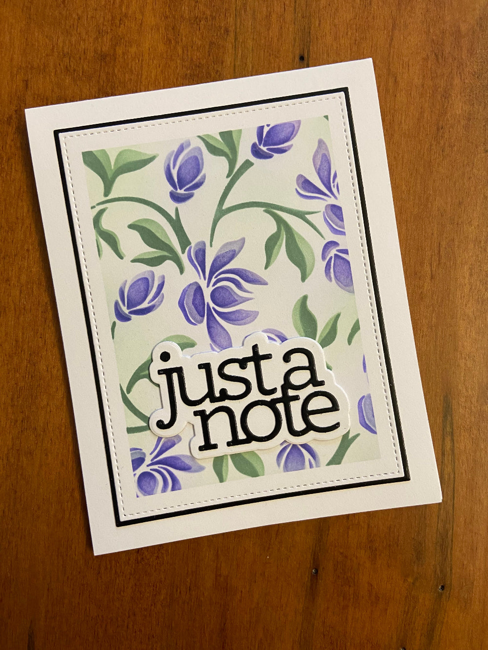 A handmade greeting card with a floral vine design in shades of purple and green on a stitched panel, with a die cut sentiment 'just a note' on a white shadow background.