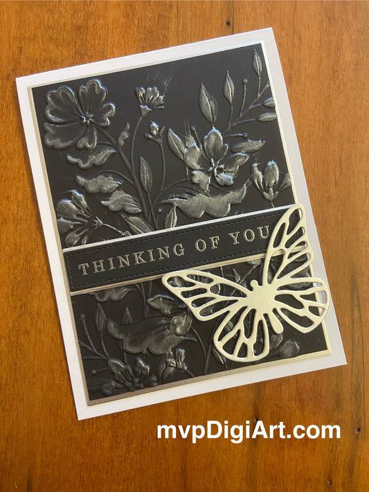 Black background with an embossed image of flowers and greenery have been lightly colourized with silver metallic ink. A thinking of you sentiment  stretches across the front. A silver die cut butterfly is attached to the bottom corner of the card. 