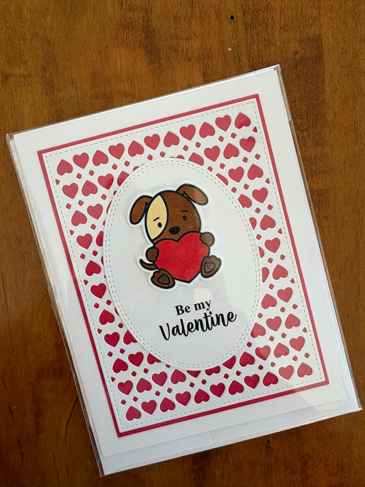Handmade Valentine's Day card featuring a sweet hand coloured dog holding up a red heart on a white oval stamped with a the sentiment "Be My Valentine". Everything is placed on top of a tiny red heart background!