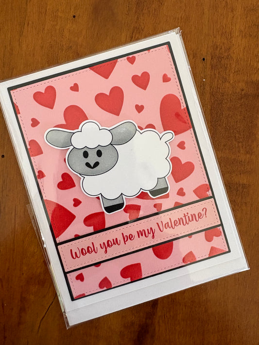 Handmade Valentine's Day card featuring an illustrated sheep surrounded by red hearts on a pink background with the pun "Wool You Be My Valentine"