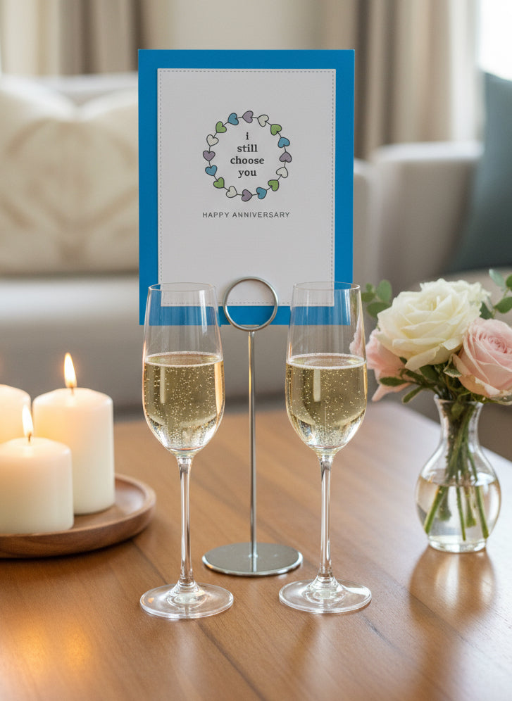 Two champagne glasses with a anniversary card on a table with candles and flowers.