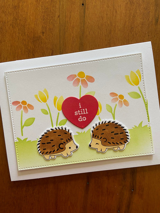 A handmade anniversary card with a hedgehog couple, flowers, and the text 'I still do' displayed on the front.