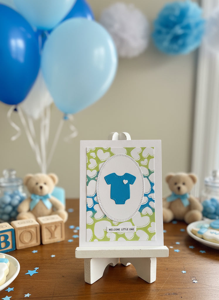 Baby shower card with blue baby onesie on a floral background, surrounded by gift boxes and balloons.