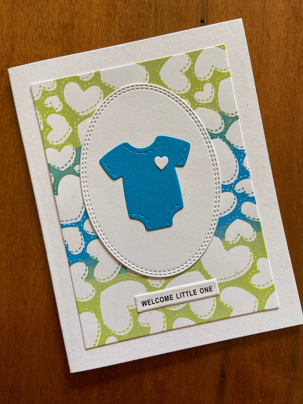 A handmade greeting card with a green and blue heart-patterned background, blue onsie and the text 'welcome little one' on a white sentiment strip.