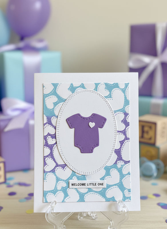 Baby shower card with purple baby onesie on a floral background, surrounded by gift boxes and balloons.