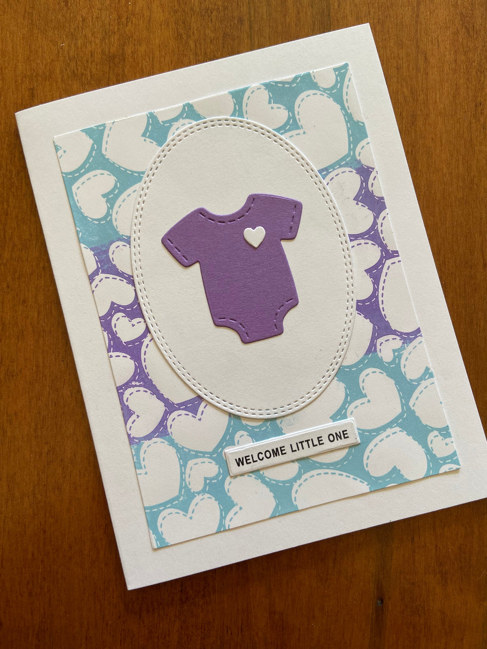A handmade greeting card with a blue and purple heart-patterned background, purple onsie and the text 'welcome little one' on a white sentiment strip.