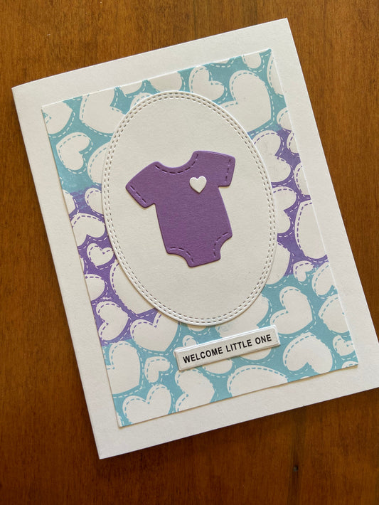 A handmade greeting card with a blue and purple heart-patterned background, purple onsie and the text 'welcome little one' on a white sentiment strip.