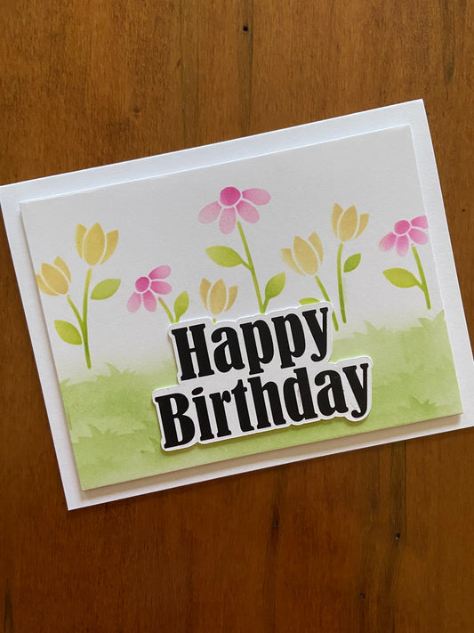 Flowers, meadow and bold happy birthday handmade card. A cute pink and yellow flowers and a grassy field scene are ink blended. A bold black happy birthday die cut sentiment tops it off. 