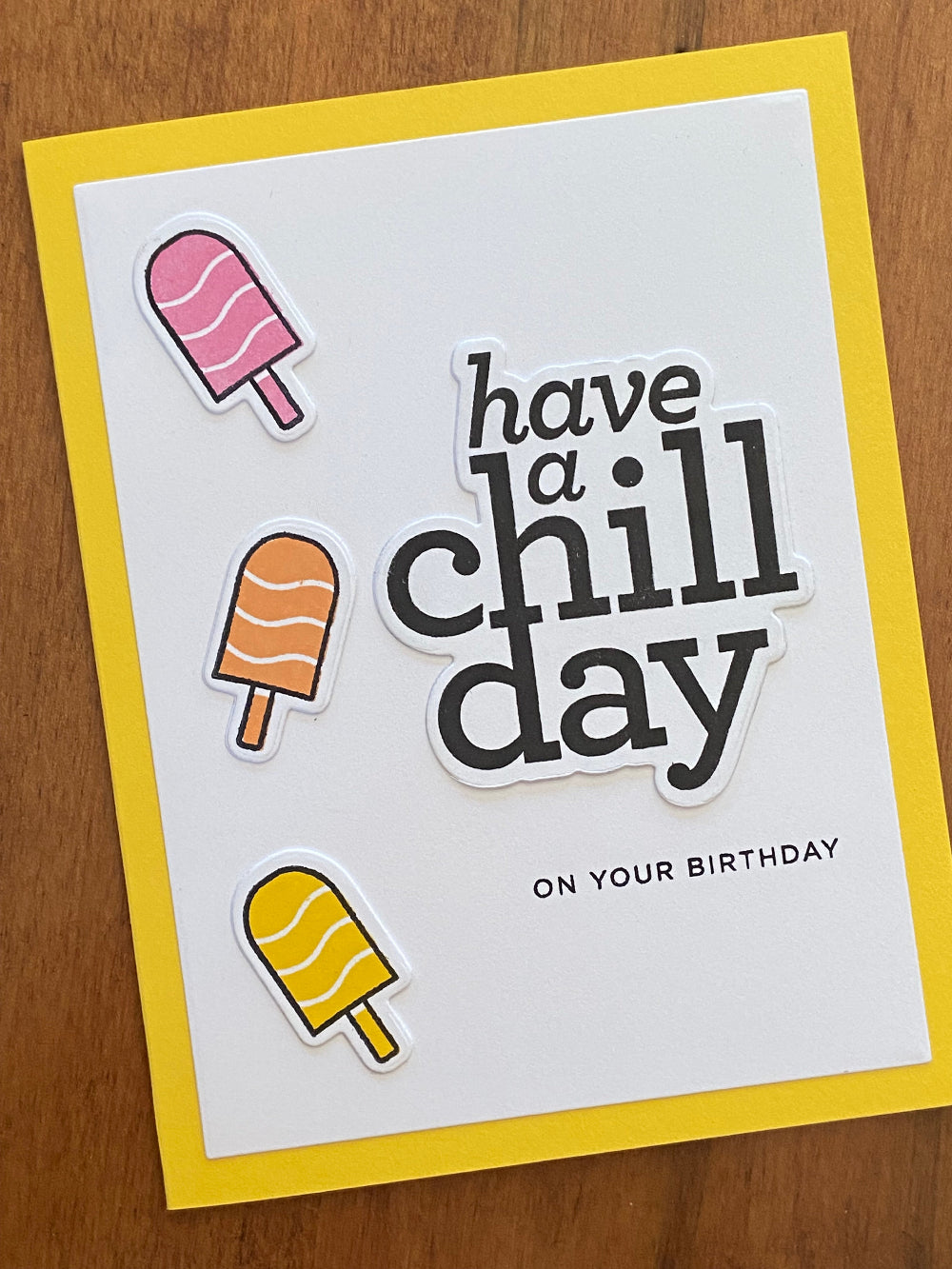 Handmade birthday card with die cut popsicles in cherry, orange, and lemon flavors, and text that reads 'have a chill day on your birthday' on a yellow notecard.