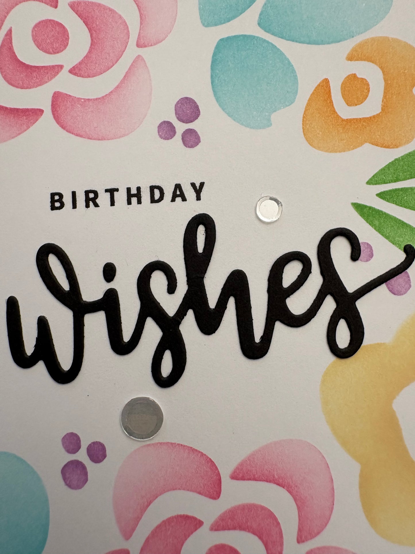 Close-up of handmade birthday card showing black die cut "Birthday Wishes" sentiment with script "wishes," stencilled pastel florals, purple ink dot clusters, and silver confetti sparkles on white cardstock