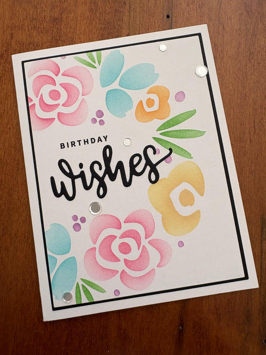 Handmade birthday card with stencilled pink roses, teal petals, and yellow flowers surrounding a black "Birthday Wishes" die cut sentiment, accented with silver confetti sparkles