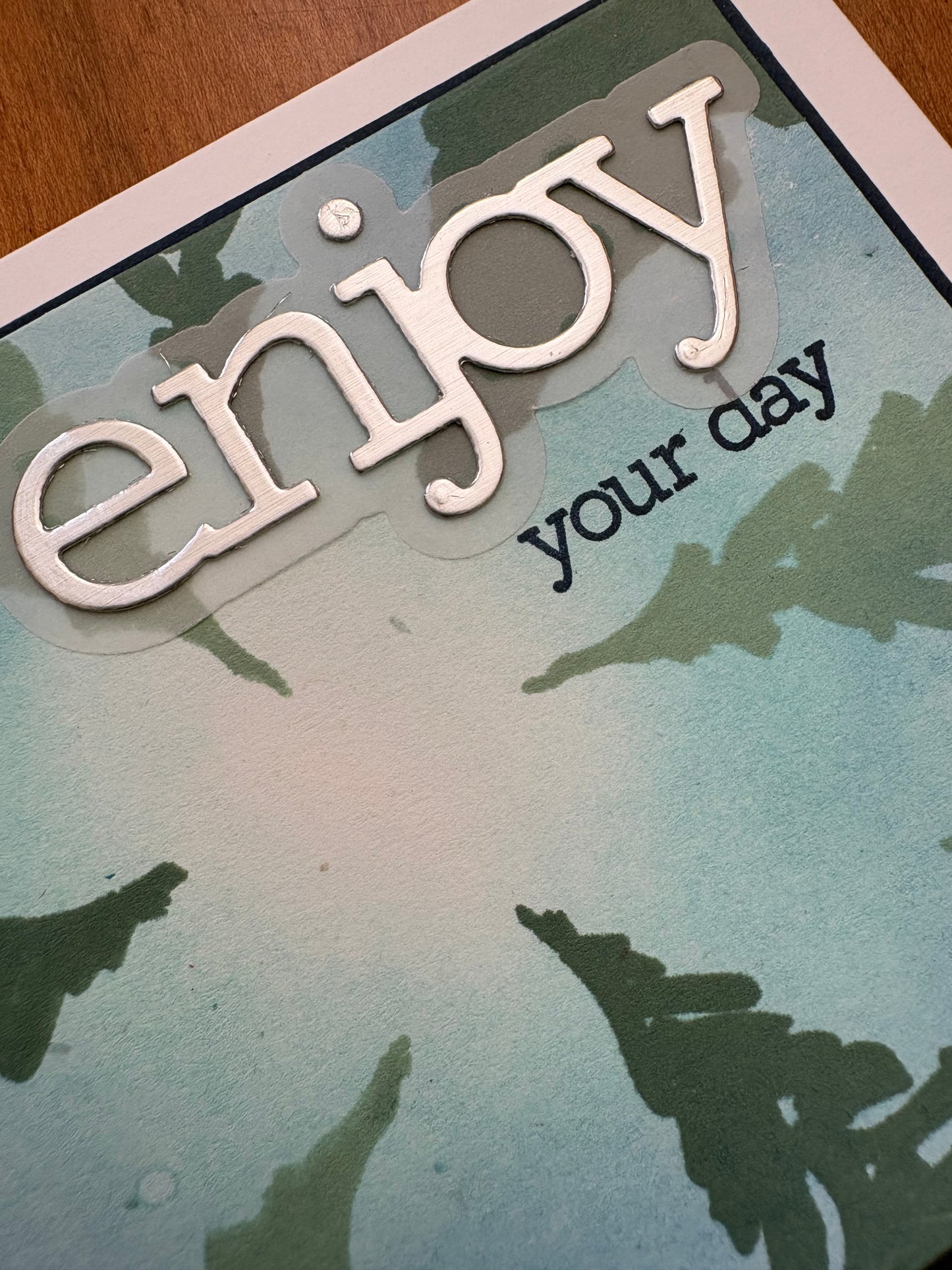 Close-up of a dimensional die cut Close up of the "enjoy" sentiment on a handmade card with ink-blended blue-green background and pine tree stencilling