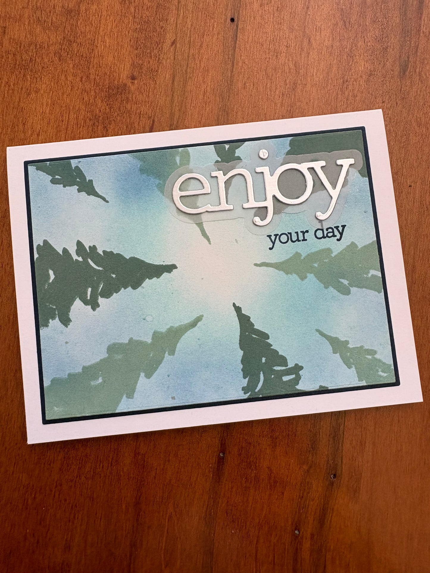 Handmade celebration card featuring a blue and green ink-blended background with stencilled pine trees and a dimensional "enjoy your day" die cut sentiment