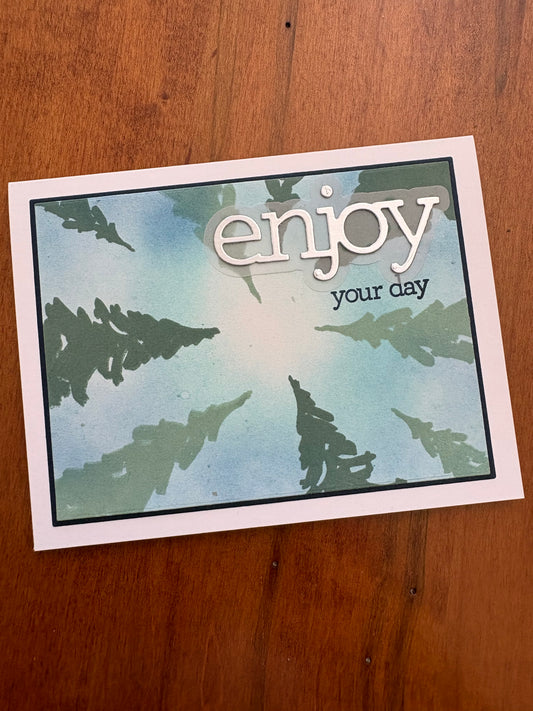 Handmade celebration card featuring a blue and green ink-blended background with stencilled pine trees and a dimensional "enjoy your day" die cut sentiment