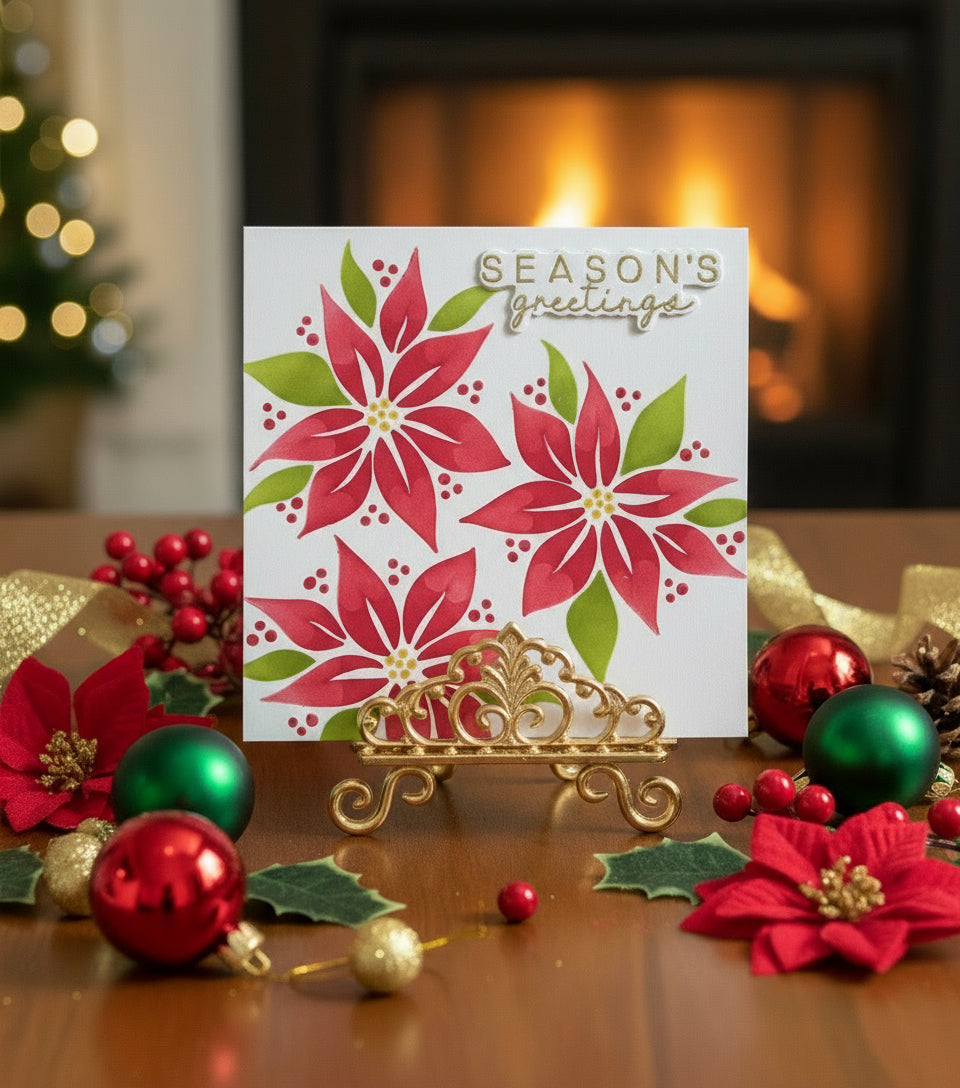 Christmas card with red poinsettias and ‘seasons greetings’ gold sentiment on a table with ornaments and a fireplace in the background,